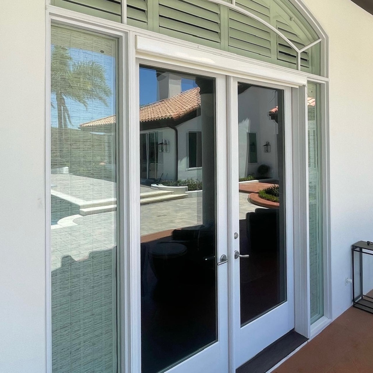 Cleaned white framed glass double doors with sidelights and a transom window reflecting a courtyard with tile roof and palm tree.