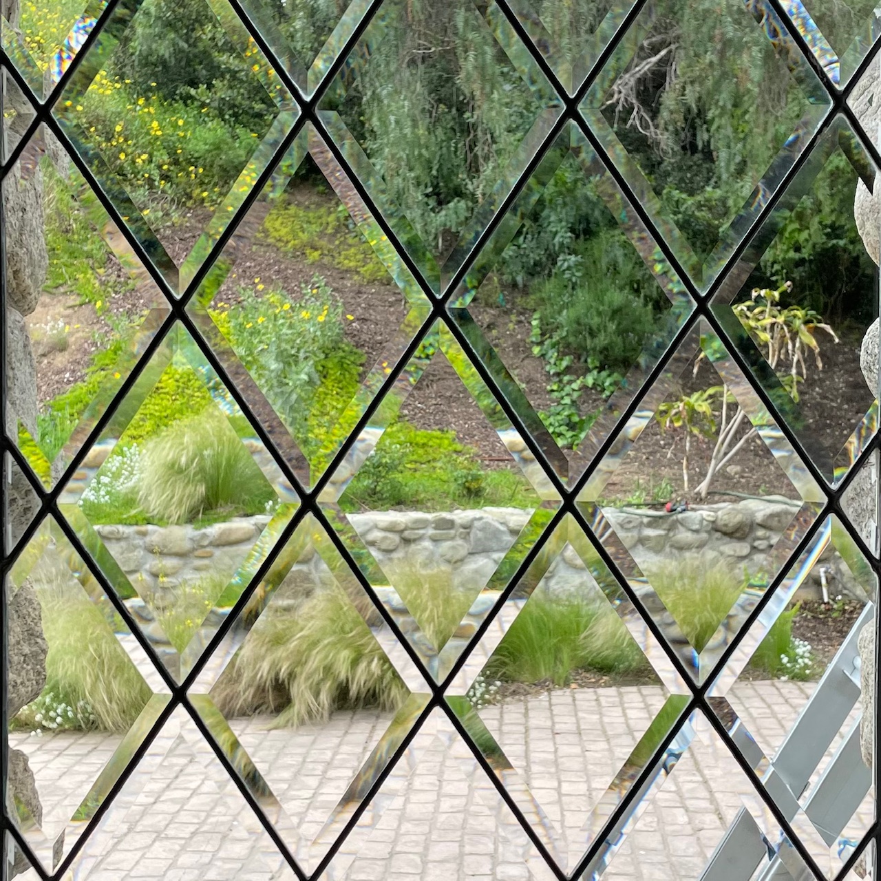 View through a cleaned diamond-patterned leaded glass window showing a stone patio, greenery, and a stone retaining wall with various plants behind it.