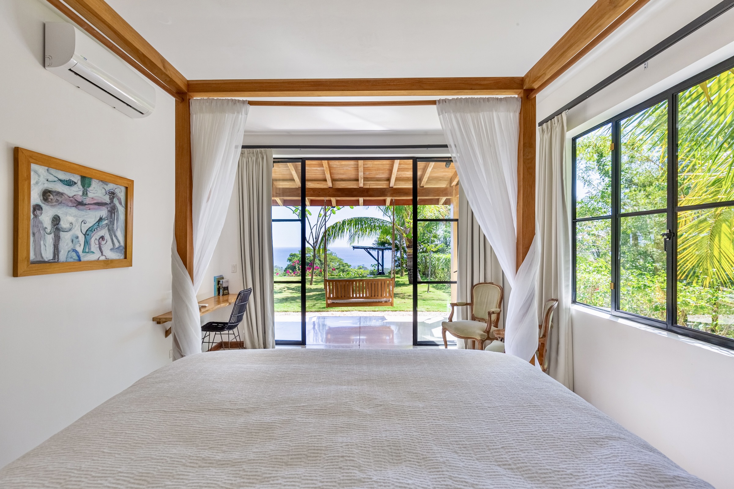 Bright bedroom with four-poster bed, sheer white curtains, and large glass doors opening to a garden with a swing and ocean view.
