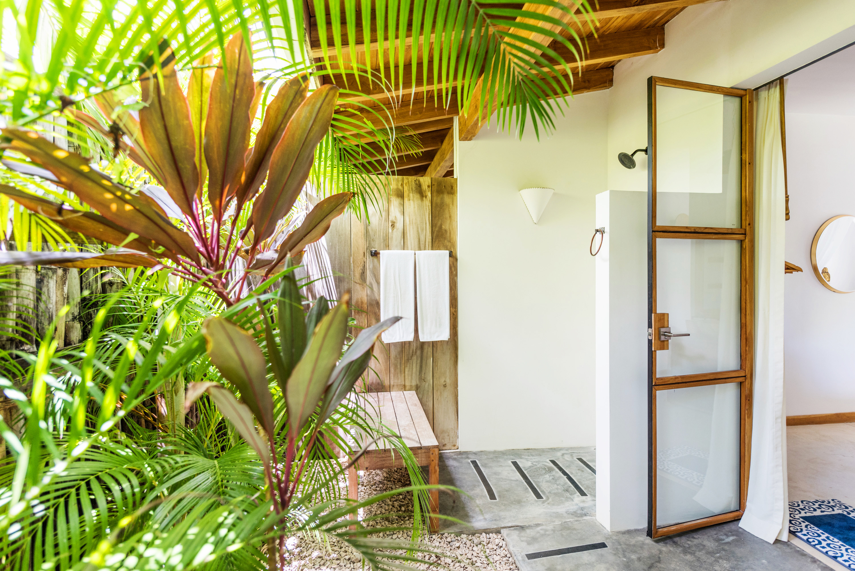 Outdoor shower area with wooden bench, two white towels hanging on a wooden wall, surrounded by lush green tropical plants and seen next to a glass door leading to an indoor space.