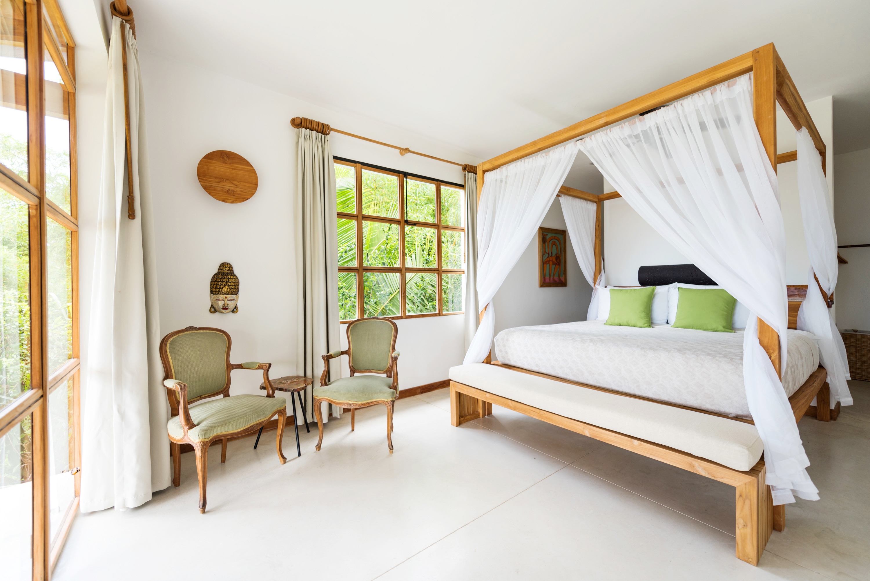 Bright bedroom with a wooden four-poster bed draped with white curtains, green pillows, two vintage chairs, and large windows with views of greenery.