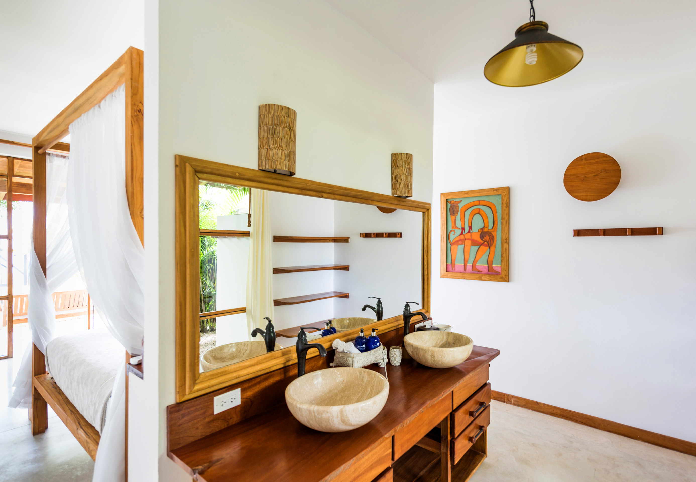 Modern bathroom vanity with two stone vessel sinks, black faucets, and a large wooden framed mirror reflecting a room with shelves and a four-poster bed draped with white curtains.