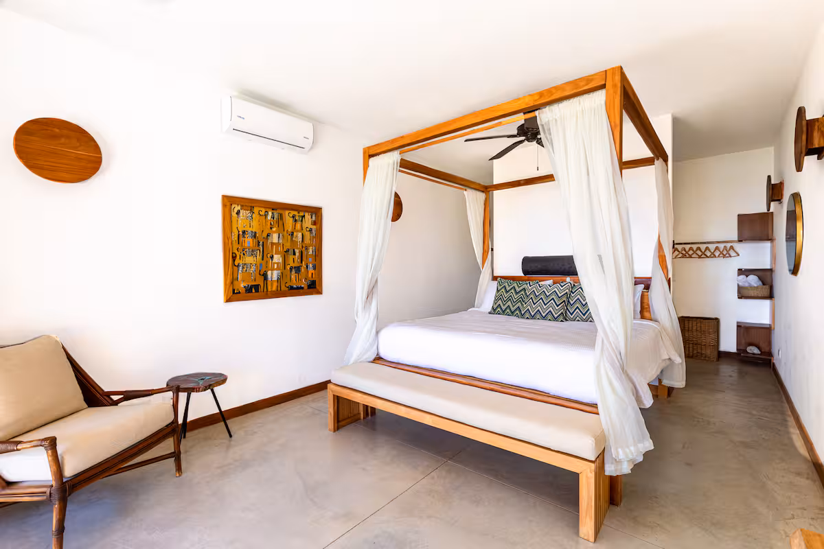 Bright bedroom with a wooden four-poster bed draped with white curtains, decorative pillows, a bench at the foot, a cushioned chair, and simple wooden wall decor.