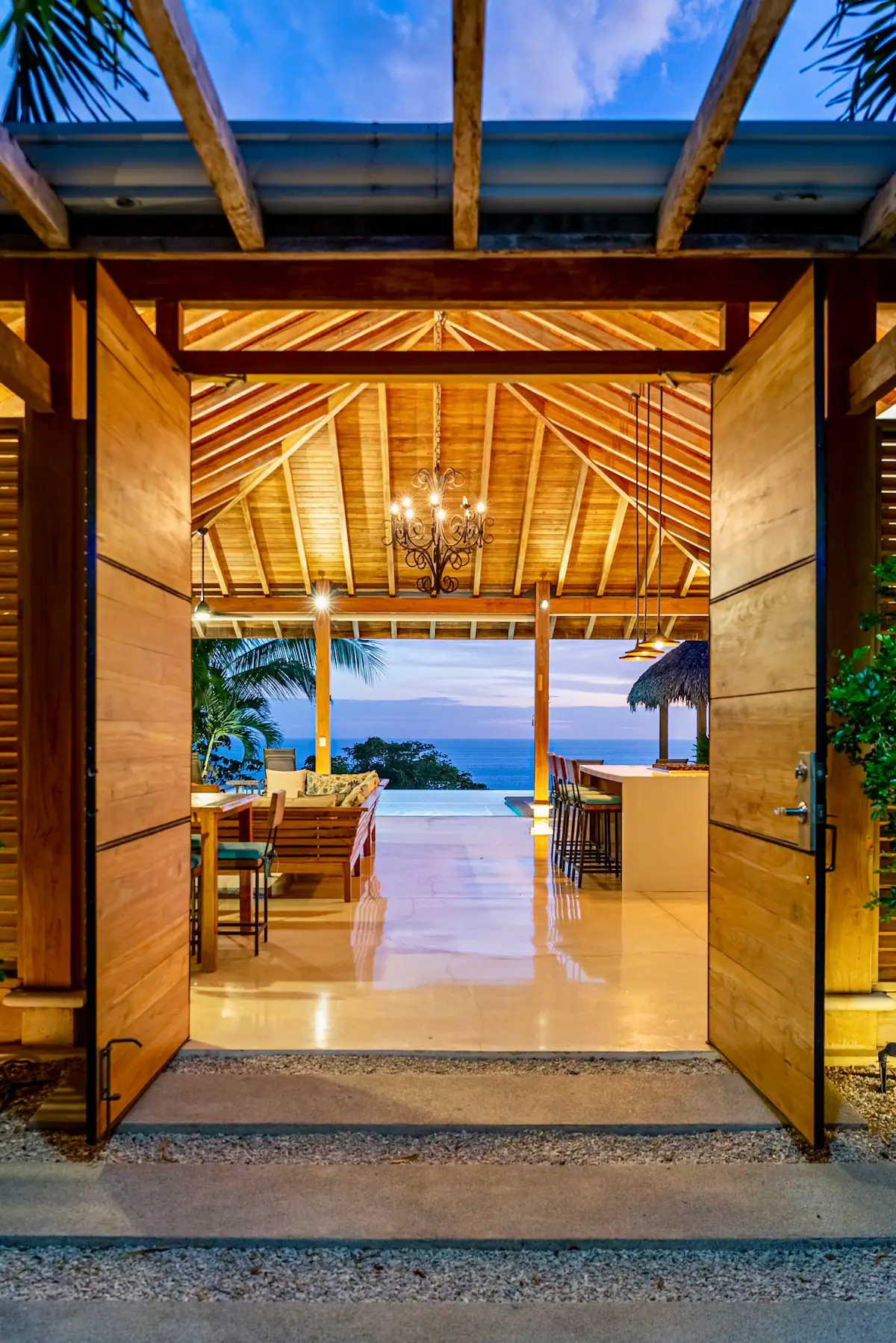 Open wooden doors leading to a spacious, illuminated patio with wooden furniture, a chandelier, and an ocean view at dusk.