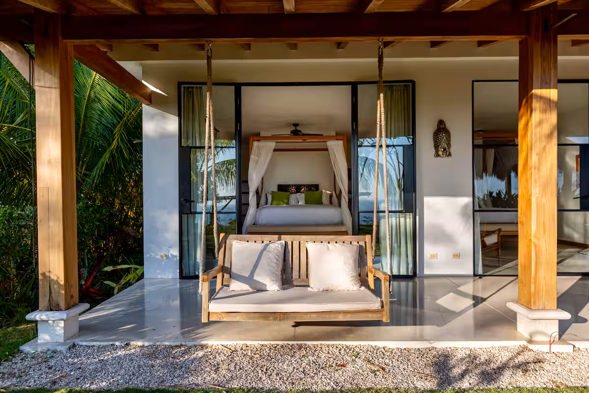 Wooden porch swing with white cushions in front of a modern bed with canopy curtains and large glass doors.
