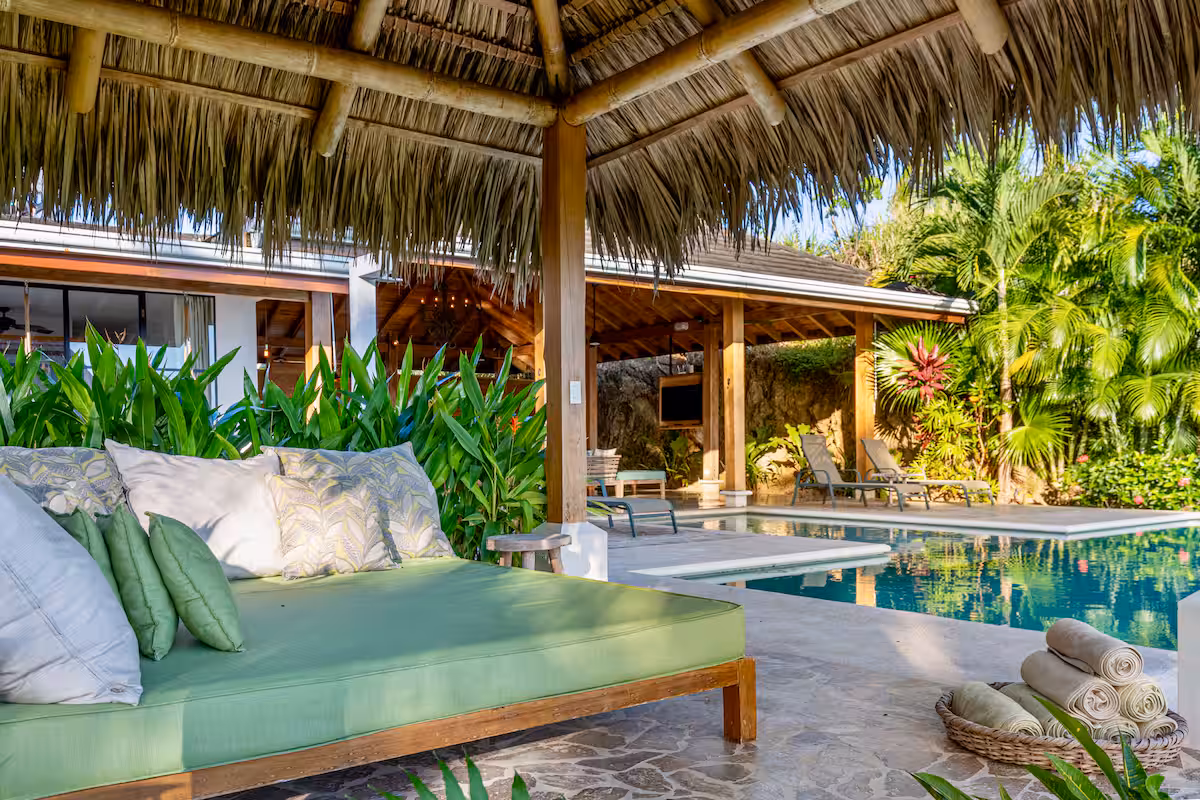 Outdoor seating area with green cushioned daybed under a thatched roof by a pool surrounded by tropical plants and lounge chairs.