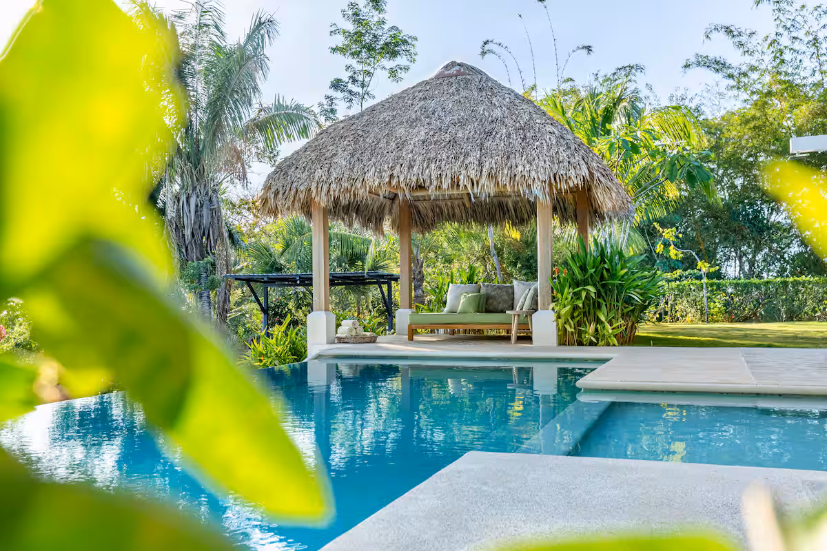 Thatched-roof gazebo with cushioned seating next to a blue swimming pool surrounded by tropical greenery.