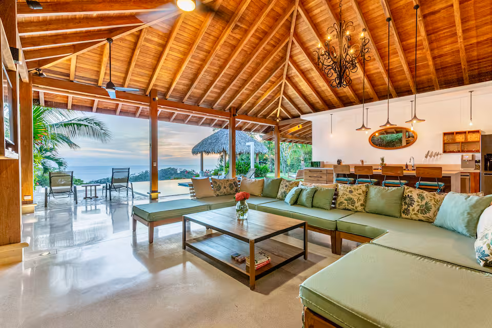 Spacious open living area with wooden vaulted ceiling, green sectional sofa, wooden coffee table, kitchen island with bar stools, and view of pool and trees outside.