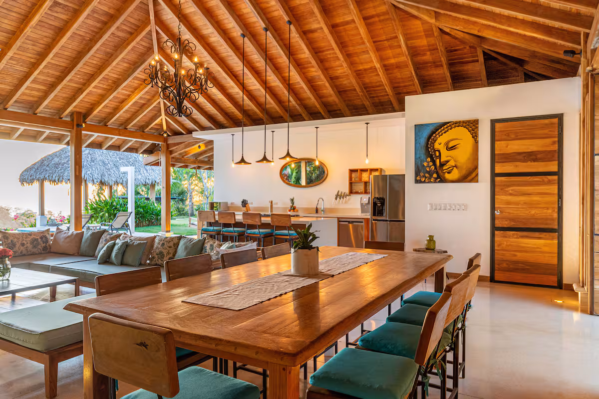 Open-concept dining and living area with a large wooden table, teal cushioned chairs, a sofa with floral cushions, and a thatched roof ceiling.