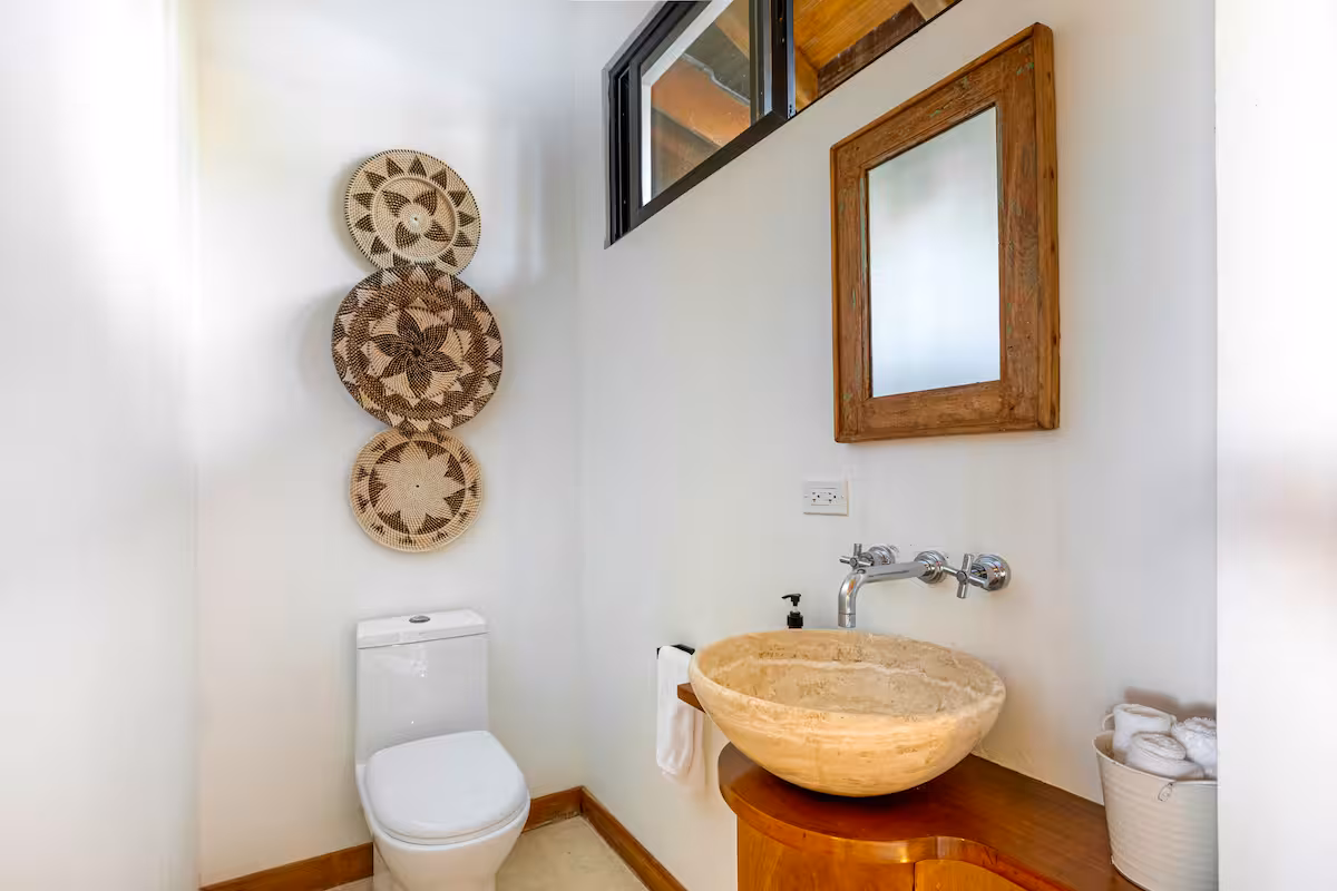 Minimalist bathroom with a white toilet, beige stone sink on wooden counter, wooden-framed mirror, and decorative woven baskets on the wall.