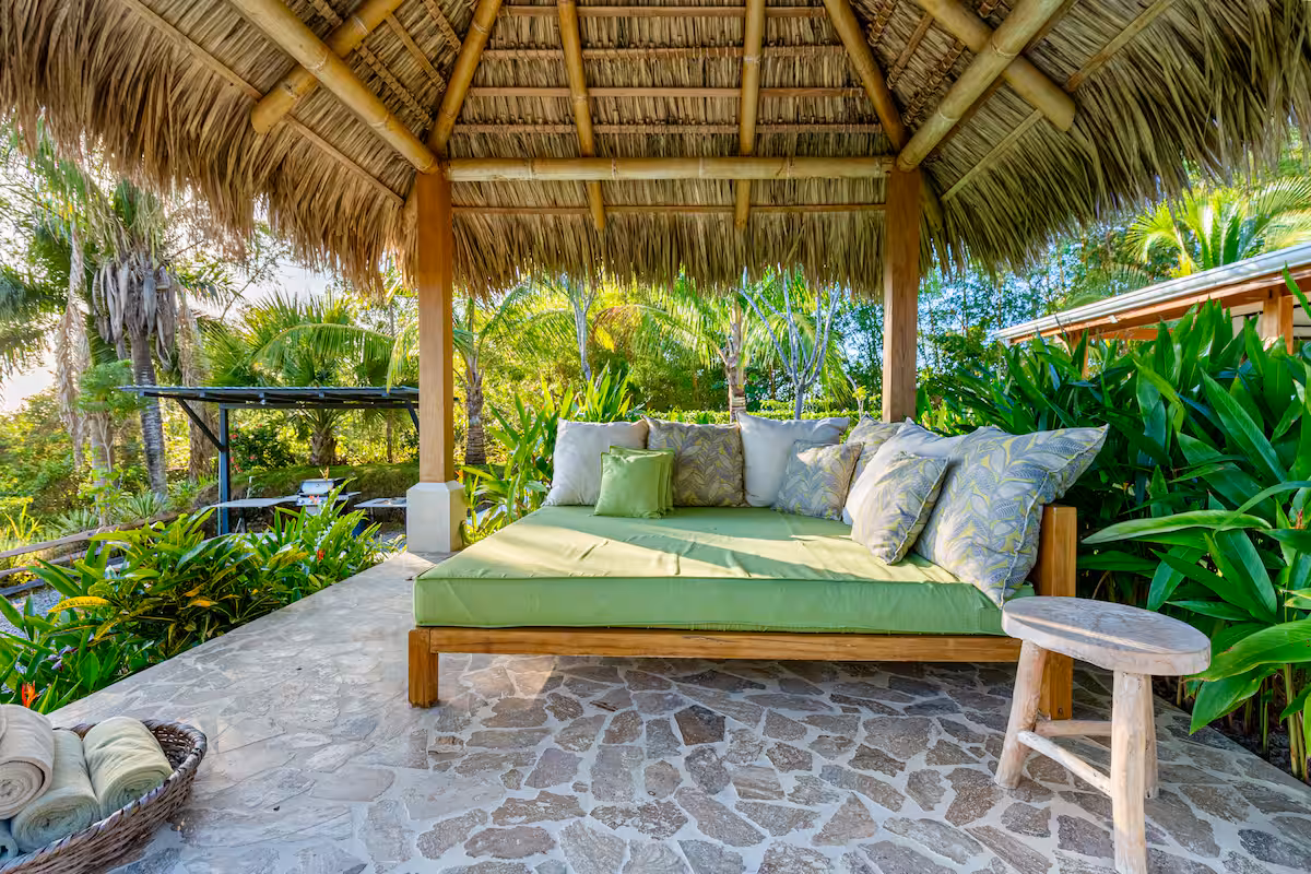 Outdoor wooden daybed with green cushions and patterned pillows under a thatched roof gazebo surrounded by lush tropical plants.