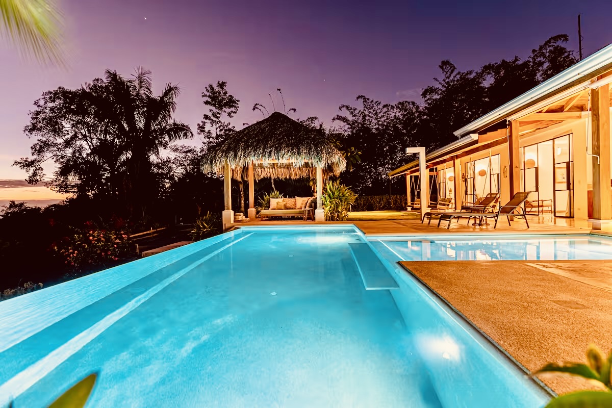 Infinity pool with lounge chairs and a thatched-roof cabana at sunset by a modern tropical house.