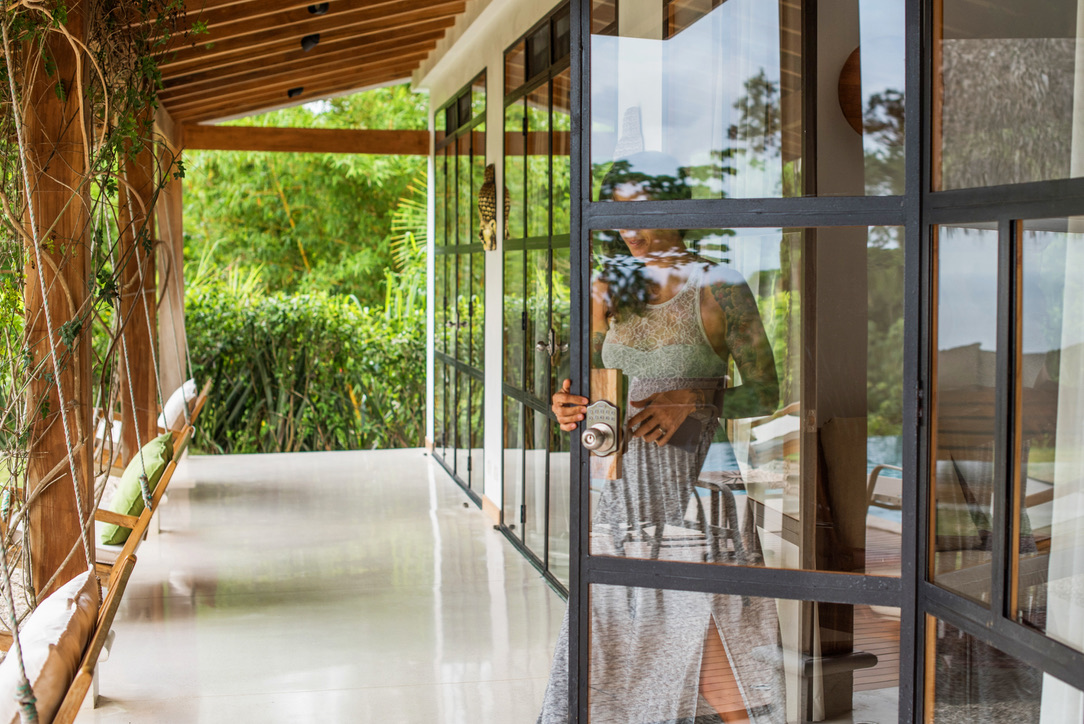 Woman in a sleeveless dress opening a glass door leading to a patio with wooden swing chairs and lush greenery.