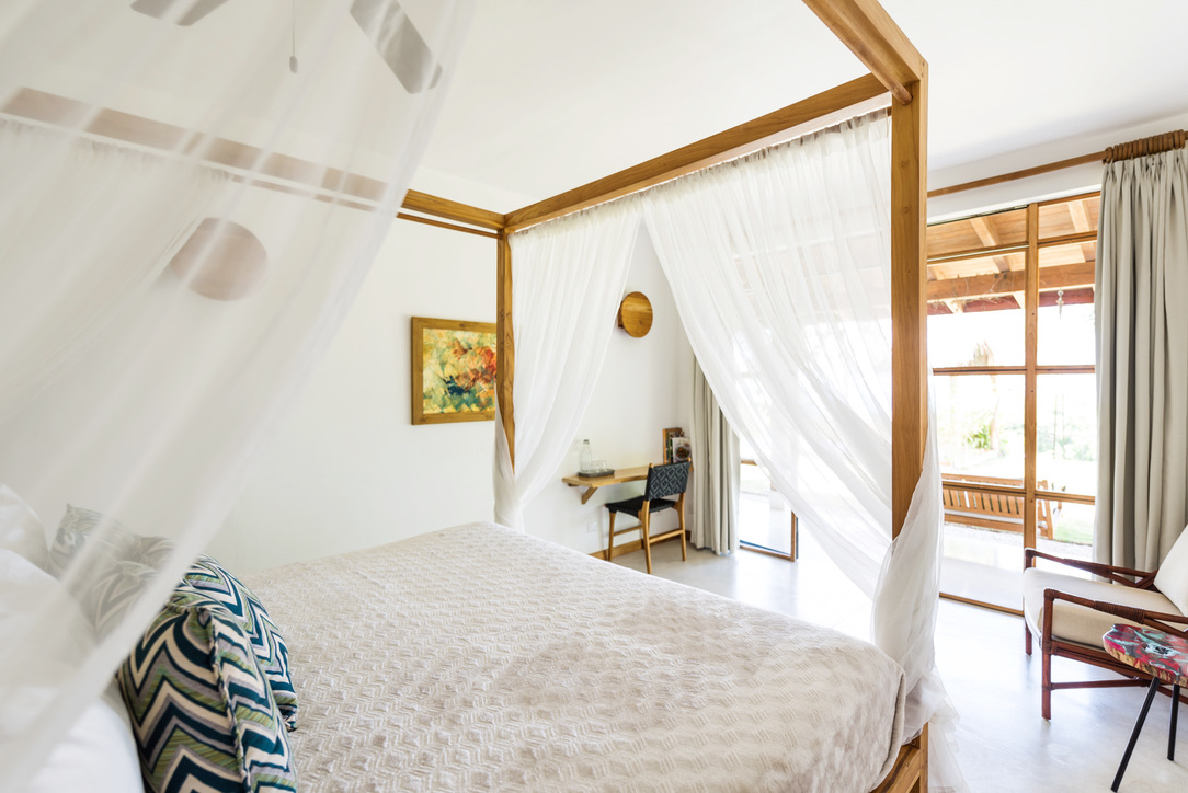Bright bedroom with a four-poster bed draped with sheer white curtains, patterned pillows, wooden desk, and sliding glass doors leading to an outdoor patio.