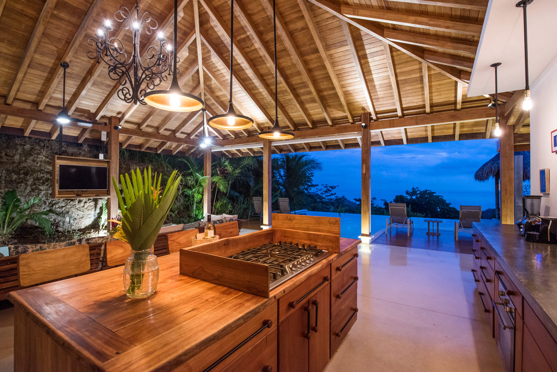 Open-air kitchen with wooden island, gas stove, hanging lights, and seating area overlooking infinity pool and ocean at dusk.