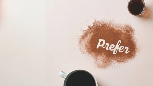 Cup of black coffee near cocoa powder spelling the word 'Prefer' on a light surface.