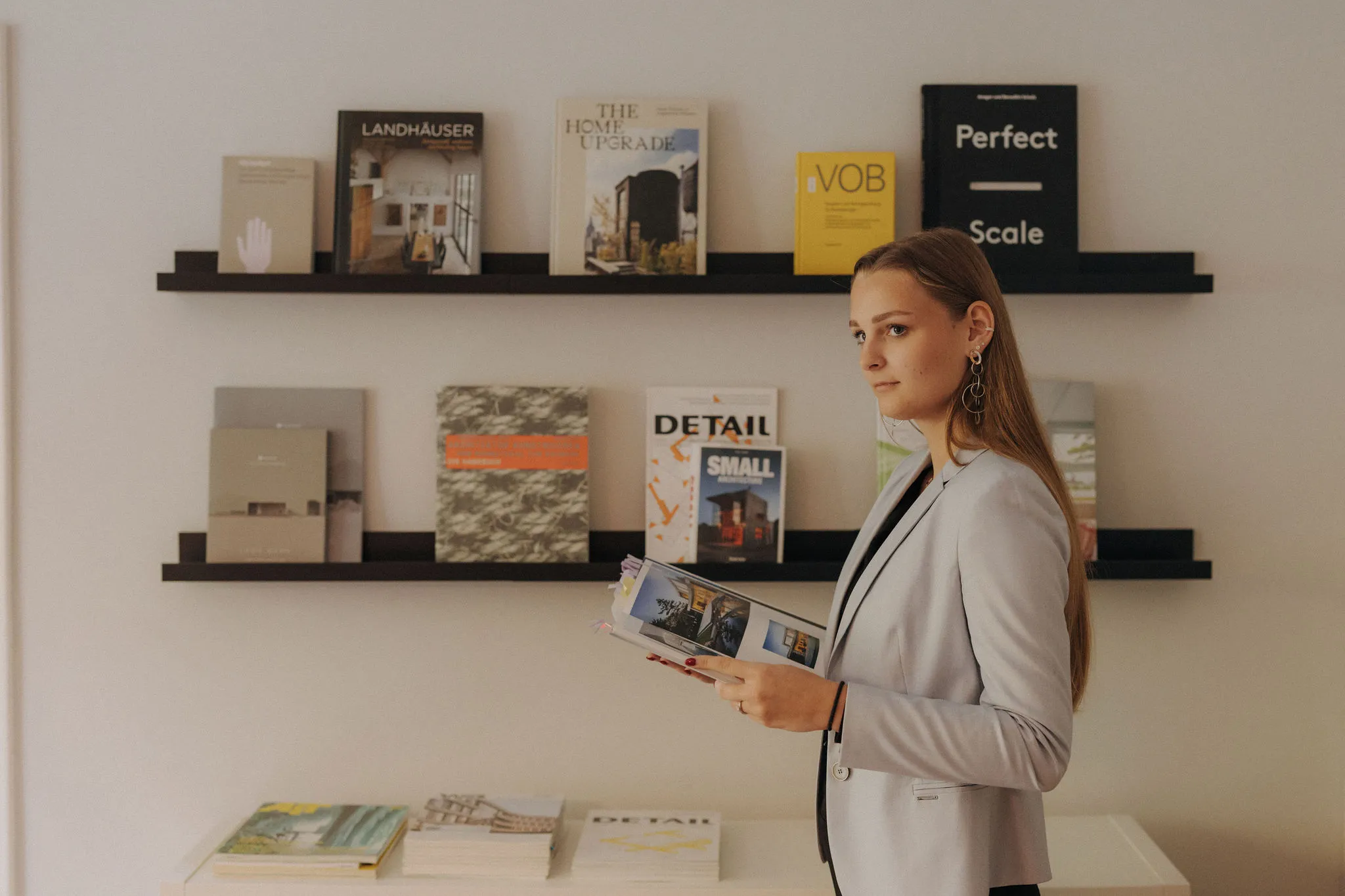 Frau mit langem Haar und grauem Blazer liest ein Magazin vor zwei Bücherregalen mit Architektur- und Designbüchern.