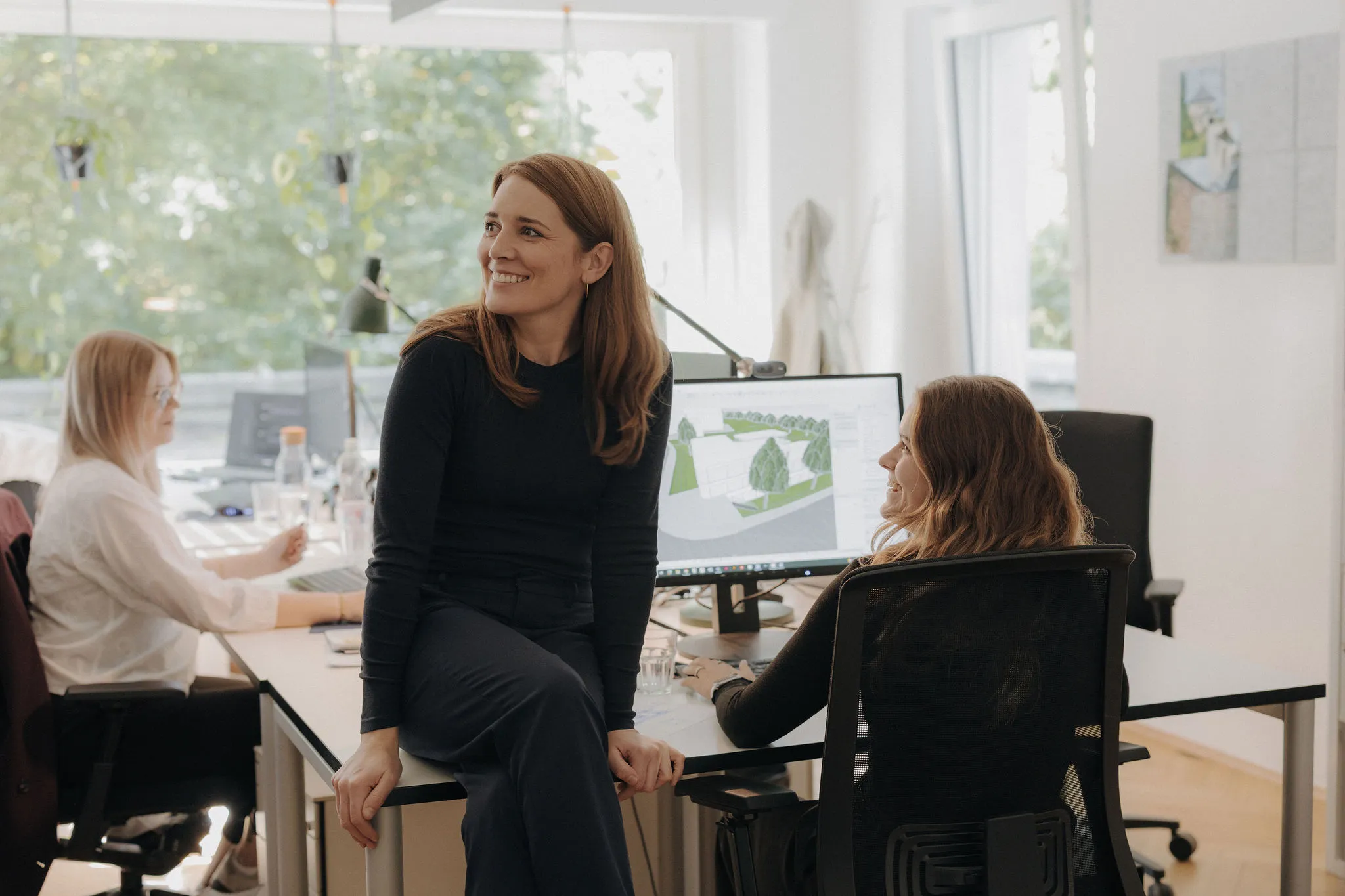 Drei Frauen arbeiten gemeinsam in einem hellen Büro mit Computern, eine sitzt auf dem Schreibtisch und lächelt.