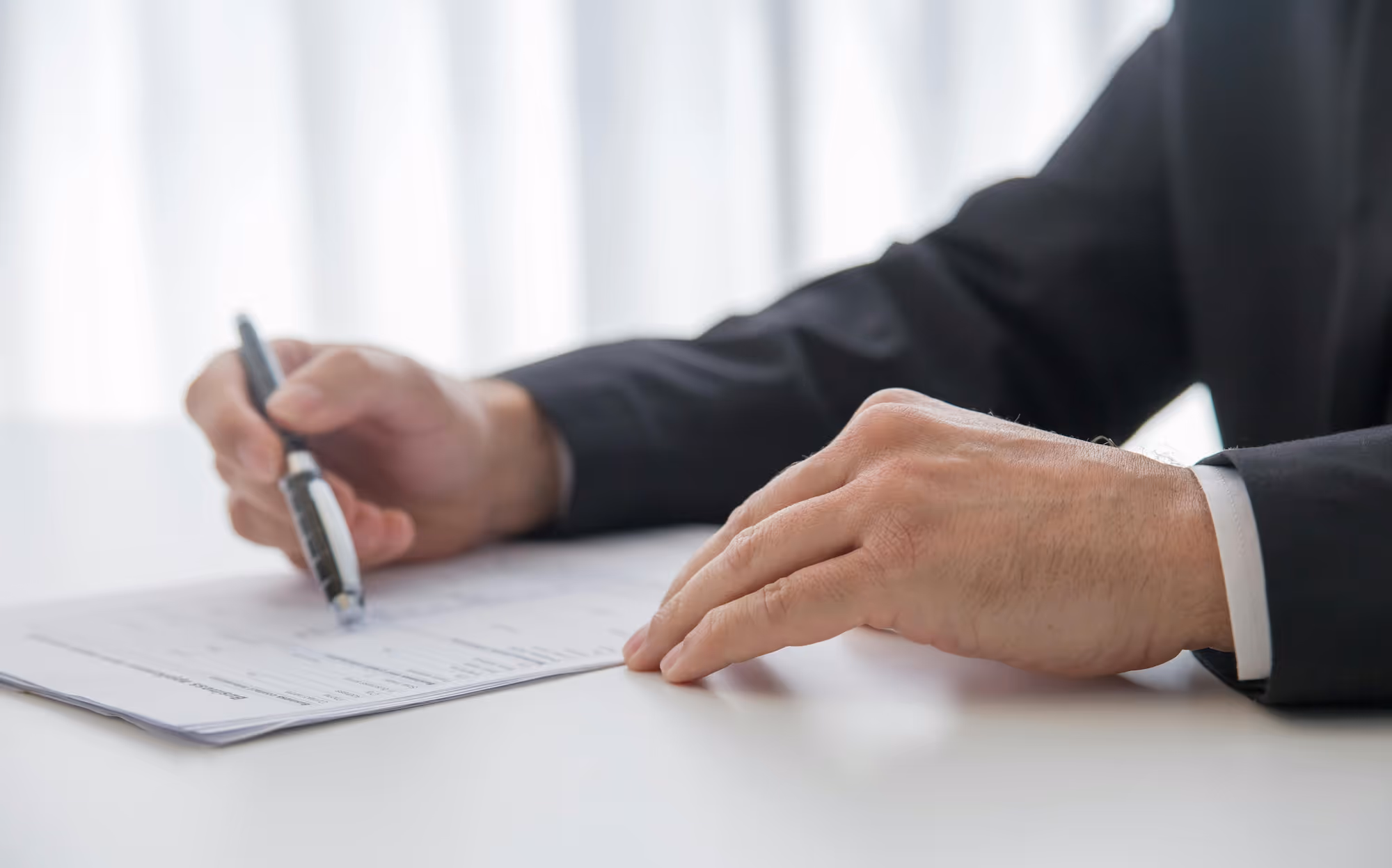 Person in a black suit writing on a document with a pen on a white table.
