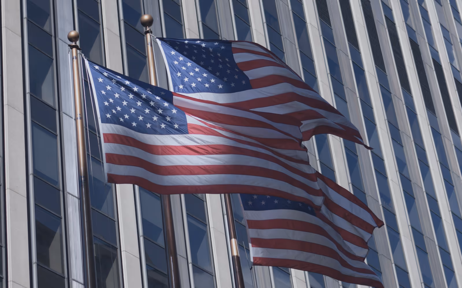 Three American flags waving on flagpoles in front of a glass and steel building.