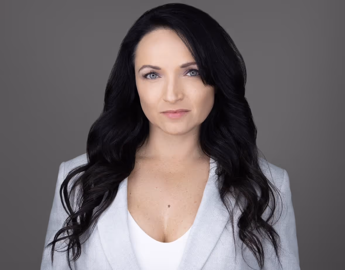 Woman with long wavy black hair wearing a light gray blazer over a white top against a neutral gray background.