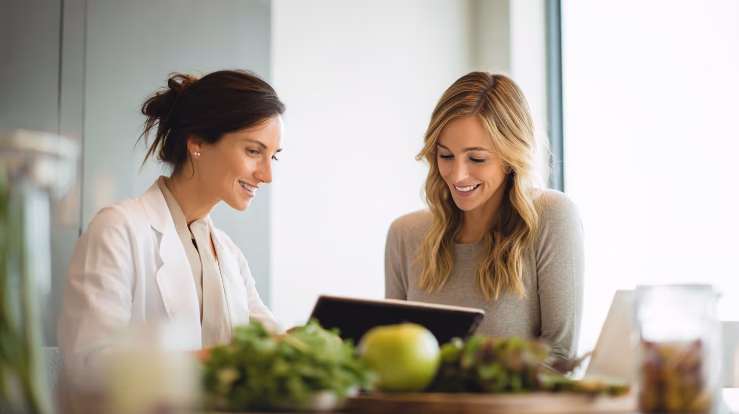 Two women smiling and looking at a tablet, with fresh fruits and vegetables in the foreground.