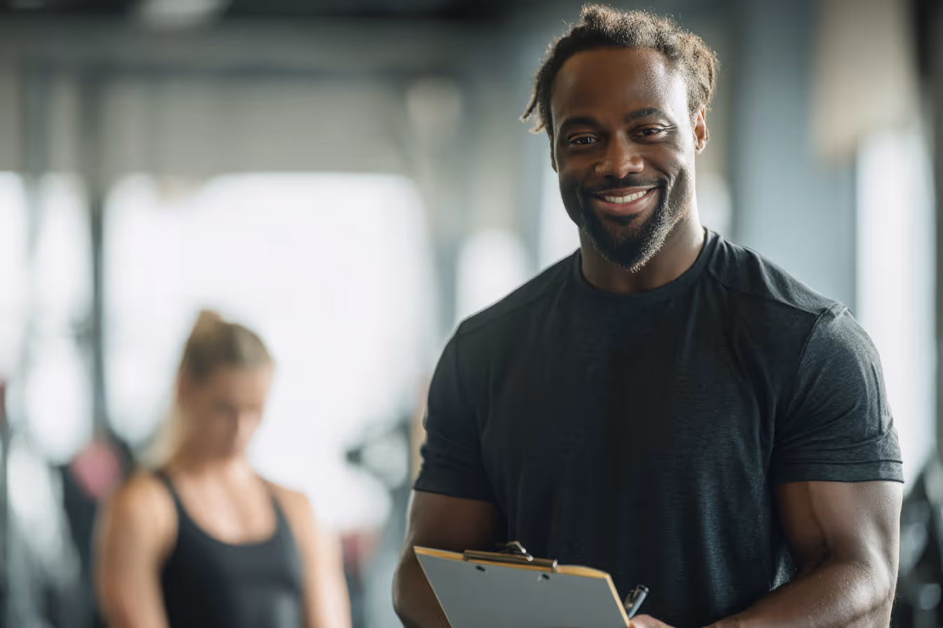 Smiling muscular man in a black t-shirt holding a clipboard in a gym with a blurred woman in workout clothes in the background.