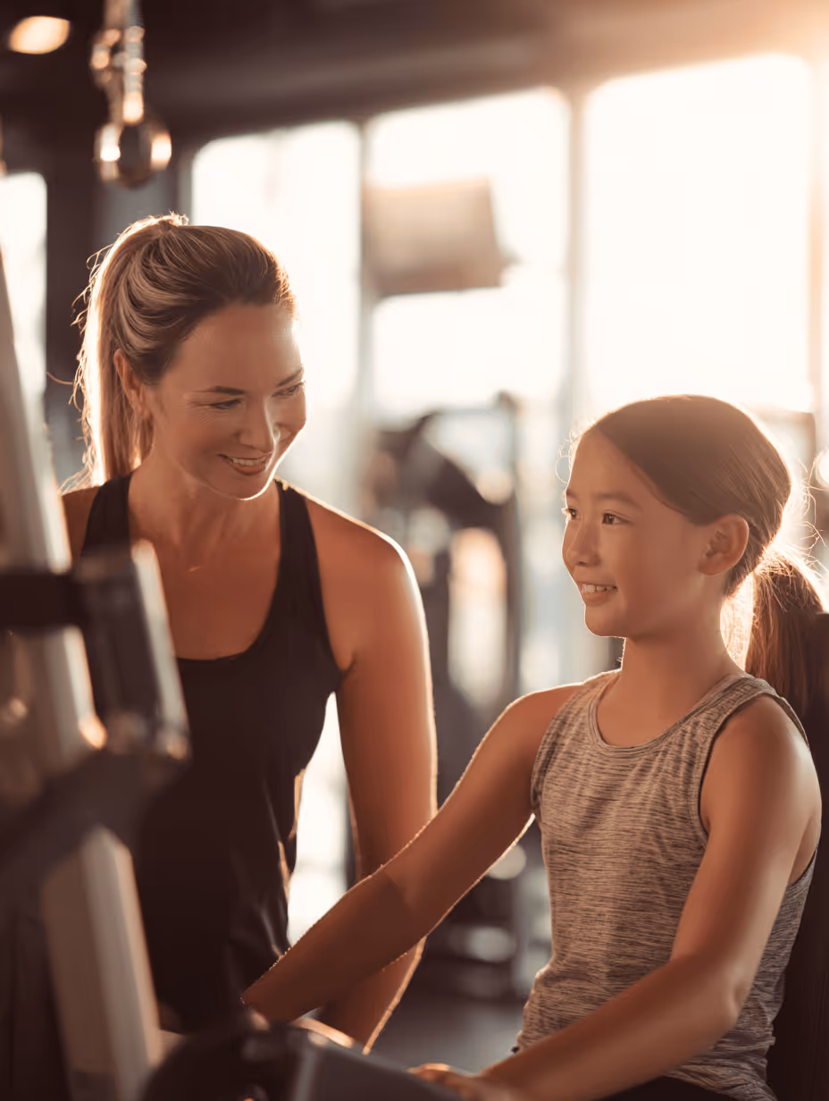 Smiling female personal trainer coaching a young girl using gym equipment with sunlight in the background.