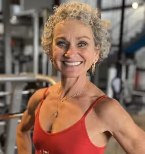 Smiling woman with curly gray hair wearing a red tank top in a gym setting.