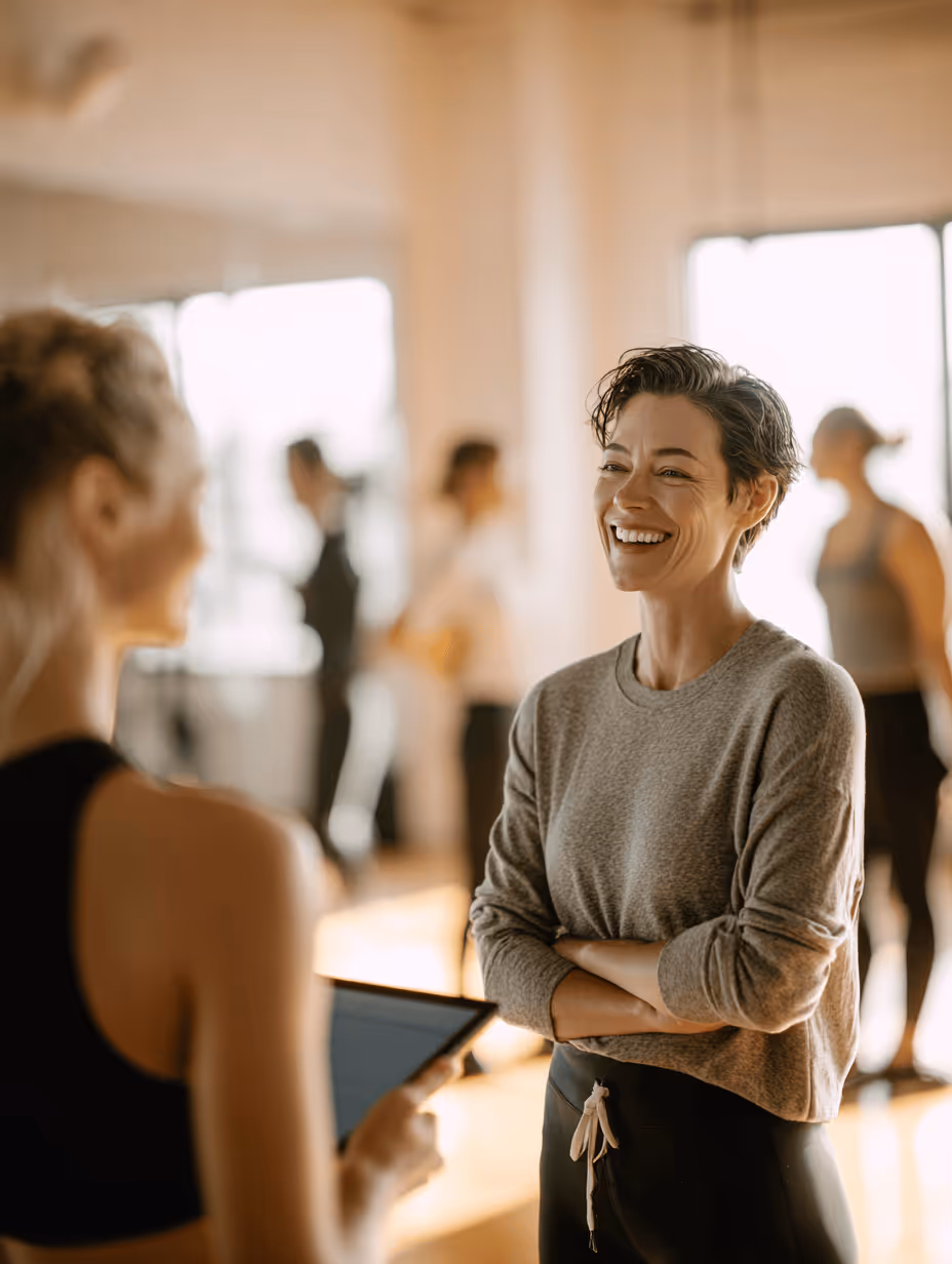Smiling woman in a gray sweater with arms crossed talking to a person holding a tablet in a bright indoor space with people in the background.