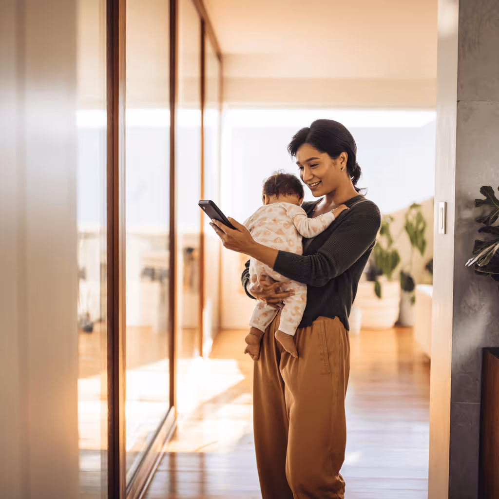 Smiling woman holding a baby and looking at a smartphone in a bright modern home.