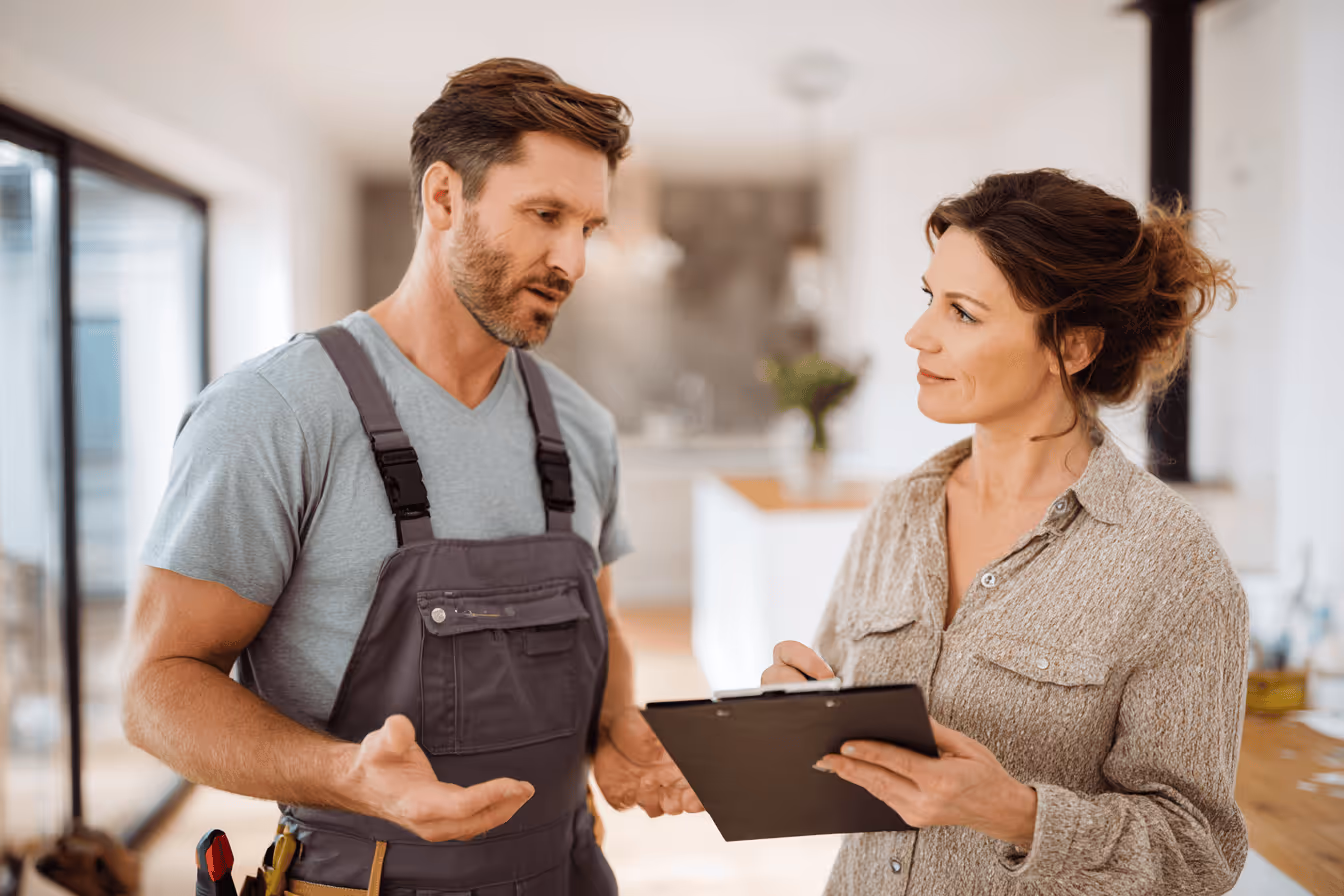 Man in work overalls discussing with woman holding a clipboard inside a modern home.