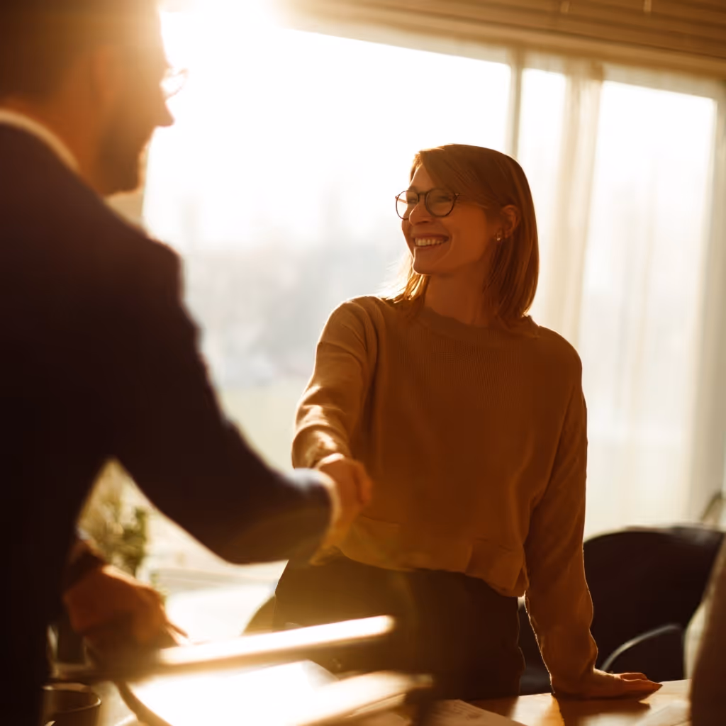 Smiling woman with glasses shaking hands with a man in a warmly lit office.