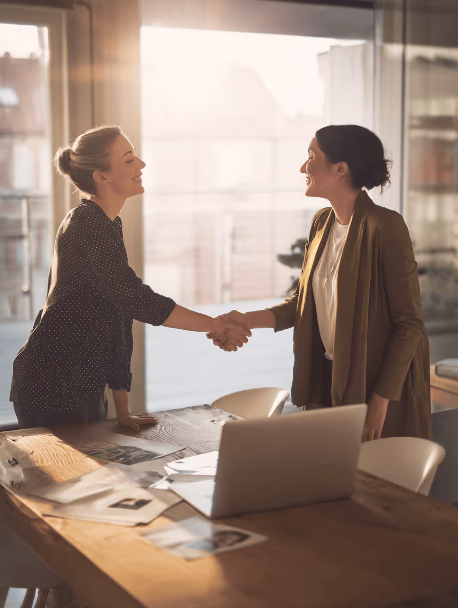 Two women smiling and shaking hands across a wooden table in a sunlit office with a laptop and documents.