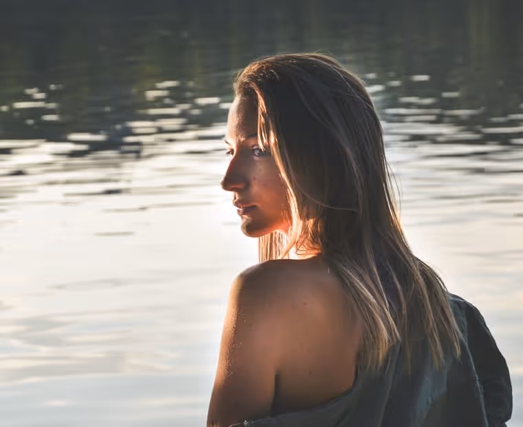 Woman with long hair wrapped in a dark towel gazing over her shoulder beside a body of calm water.