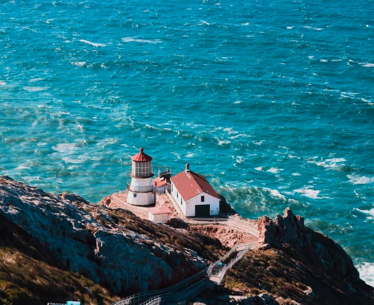 Lighthouse with red roof and white walls situated on rocky cliffs overlooking turbulent turquoise ocean waters.