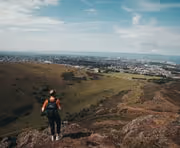 Person hiking on rocky terrain overlooking a green valley with a distant town under a blue sky with clouds.