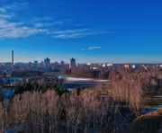 City skyline with a mix of tall buildings and smokestacks under a blue sky, with leafless trees and a water body in the foreground.