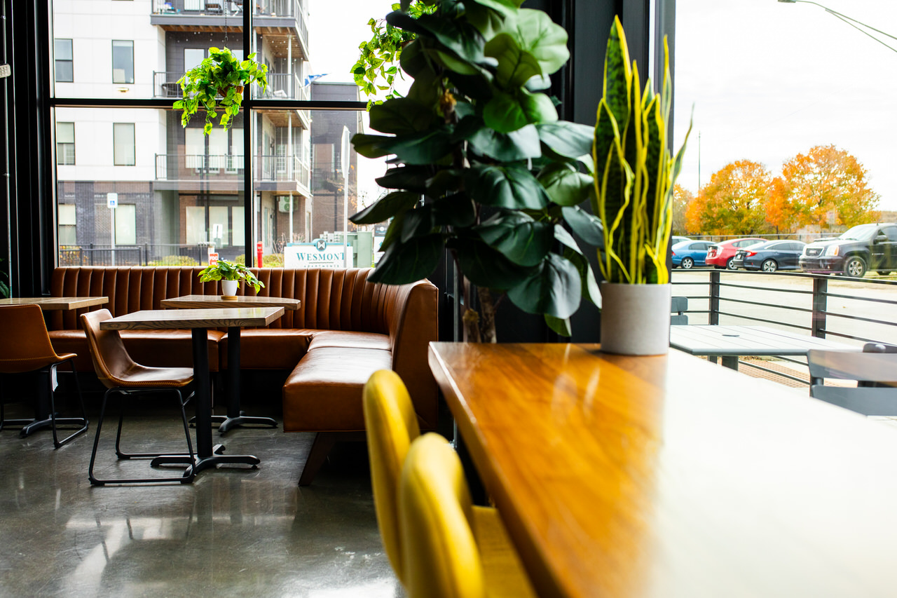Green Room - Modern café interior with brown leather booth seating, wooden tables, indoor plants, and large windows showing parked cars outside.