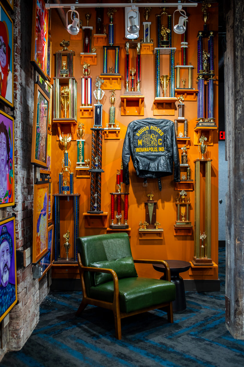 Polk Micro Museum - Green leather armchair with pillow in front of a wall displaying numerous trophies and a black leather jacket with 'Rough Riders MC Indianapolis, Ind.' in yellow lettering.