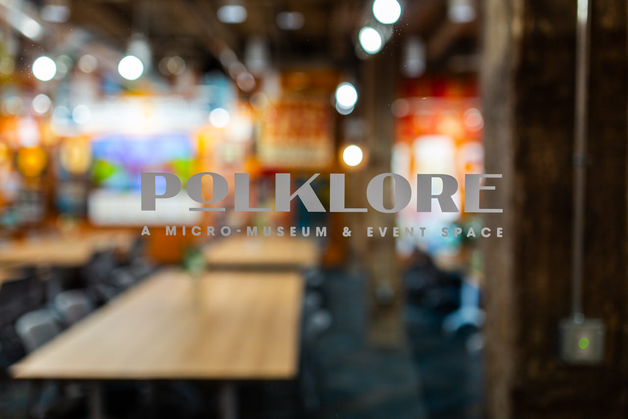 POLKLORE - Blurry interior view of a micro-museum and event space with wooden tables and chairs behind glass with the text 'POLKLORE A MICRO-MUSEUM & EVENT SPACE'.