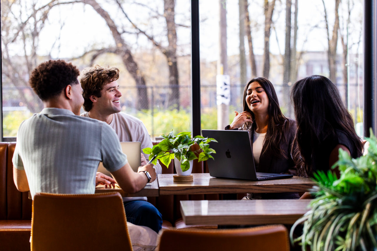 Four young adults sitting at a wooden table with laptops, smiling and conversing in a bright room with large windows and green plants.