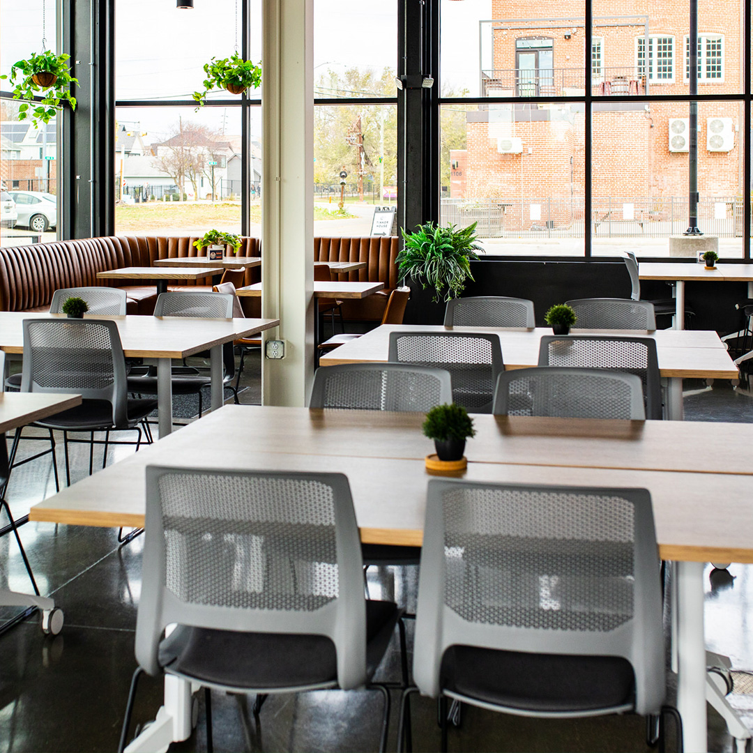 Modern coworking space with wooden tables, gray mesh chairs, potted plants on each table, and large windows showing a brick building outside.