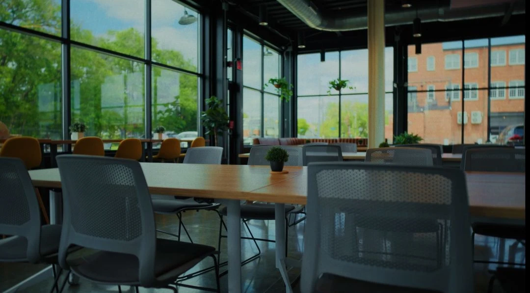 Modern coworking space with large windows, wooden tables, gray and yellow chairs, and potted plants.