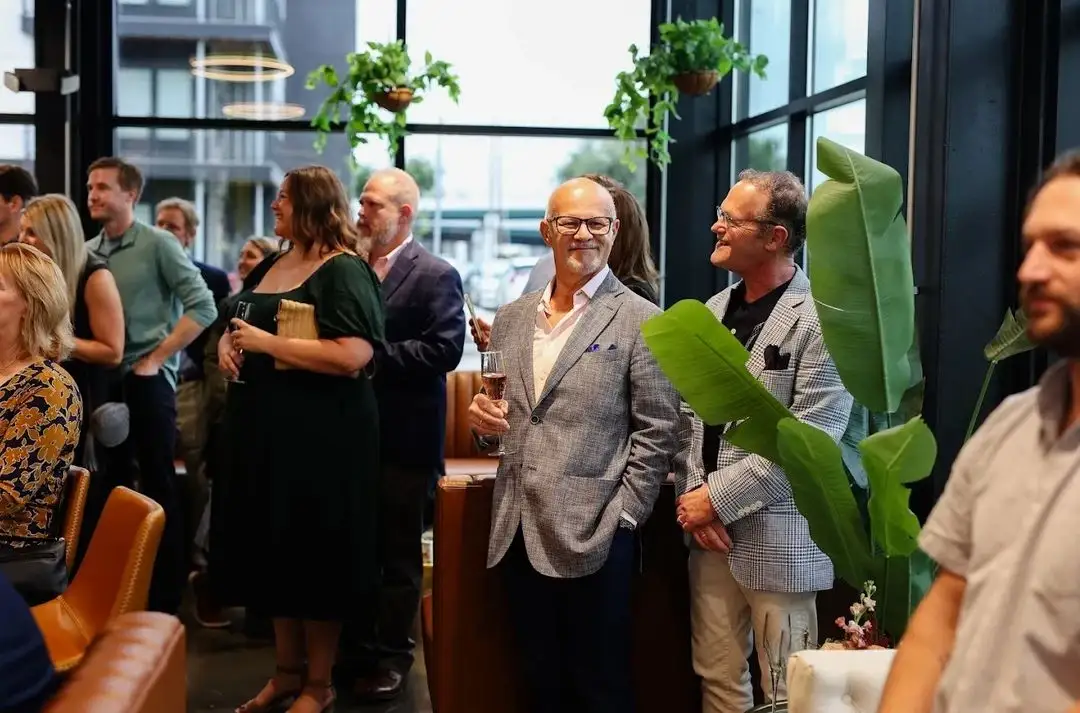 People dressed in smart casual attire socializing in a modern indoor space with large windows and plants.