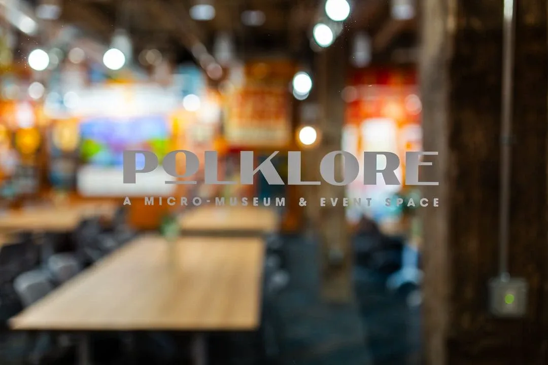 Glass door with POLKLORE micro-museum and event space logo, blurred interior with tables and chairs behind it.