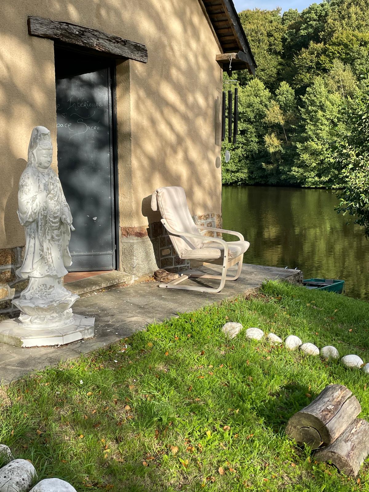 Statue blanche devant une porte noire à côté d'un fauteuil beige sur un patio, avec un lac et des arbres en arrière-plan.