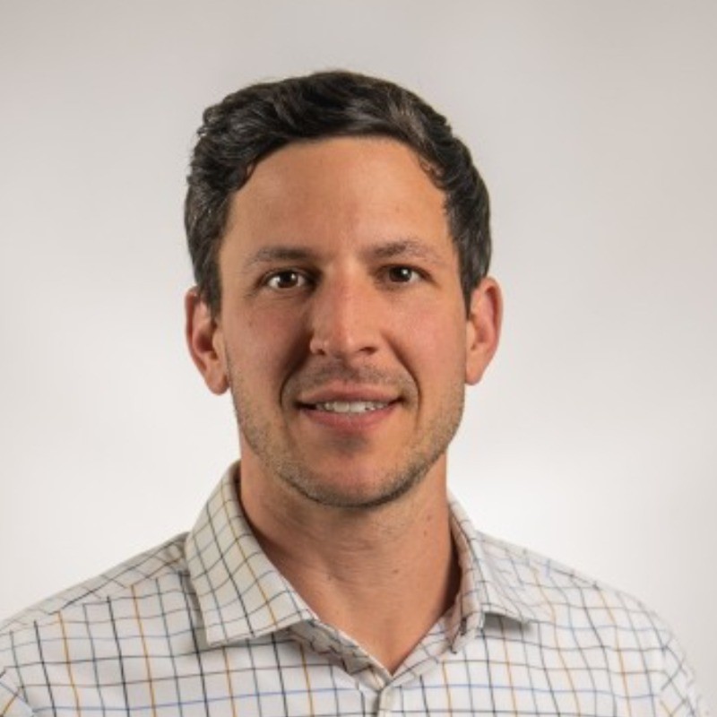 Man with short curly dark hair wearing a white checkered button-up shirt smiling against a plain light background.