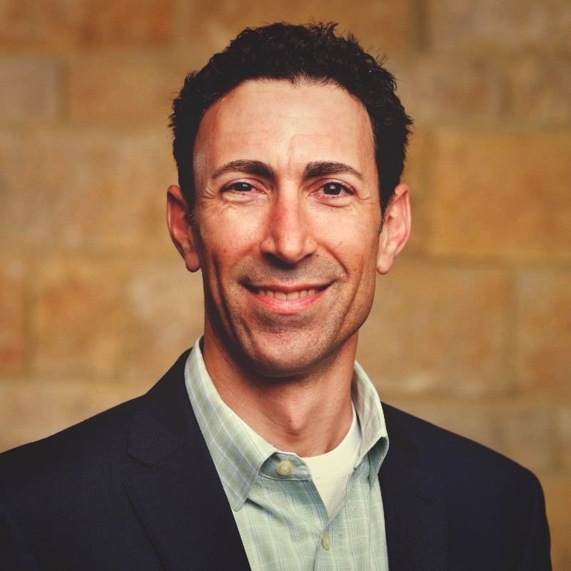 Smiling man with curly dark hair wearing a dark blazer and green checkered shirt against a blurred brown brick background.
