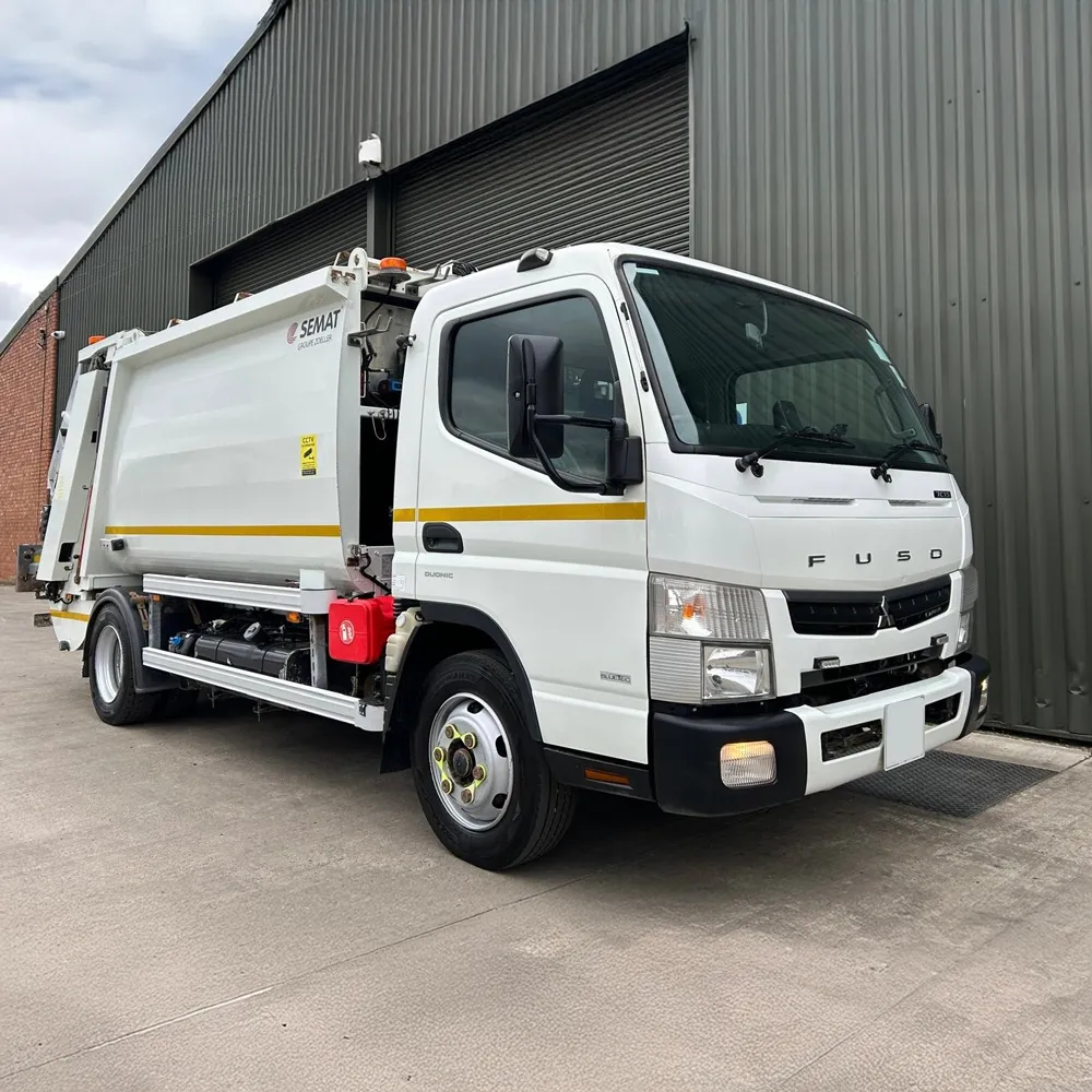 White Fuso refuse collection truck with yellow stripe parked outside a warehouse with gray metal siding.